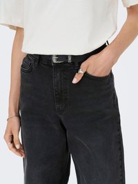 Frau trägt schwarze High-Waist-Jeans mit einem dekorativen schwarzen Gürtel, weißem Oberteil und silbernem Schmuck, eine Hand in der Tasche.
