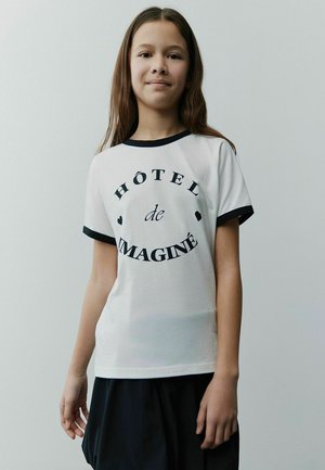 Weißes T-Shirt mit schwarzem Saum, mit "HÔTEL de IMAGINÉ" in fettem Text bedruckt, kurze Ärmel und lockere Passform, getragen mit einem dunklen Rock.