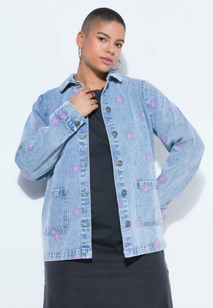 Osoba s krátkými vlasy, která má na sobě kruhové náušnice a džínovou bundu s růžovými slunečními vzory přes černé šaty, pózující na bílém pozadí.