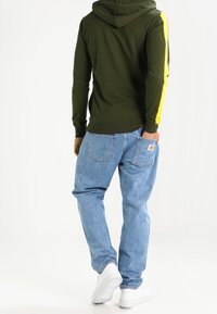 Uomo che indossa una felpa verde oliva con strisce gialle sulle maniche, jeans azzurri chiari e scarpe da ginnastica bianche, in piedi con la schiena rivolta in avanti.