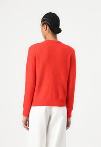 Red cashmere sweater with long sleeves, ribbed cuffs and hem, smooth texture, and rounded neckline. Worn with light-colored pants.