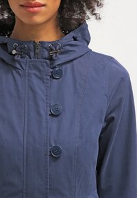 Tummansininen vedenpitävä takki, jossa on huppu, etuvetoketju ja suuret napin korostukset. Sujuva texture ja hienovarainen kiilto.