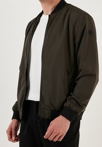 Oliivinvihreä pommikankainen takki, jonka materiaali on sileä. Takissa on vetoketju edessä, resorit hihansuissa ja kaksi sivutaskua.