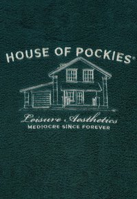 Zielony ręcznik z białym, haftowanym napisem "HOUSE OF POCKIES" nad grafiką domu oraz "Leisure Aesthetics Mediocre Since Forever" poniżej.