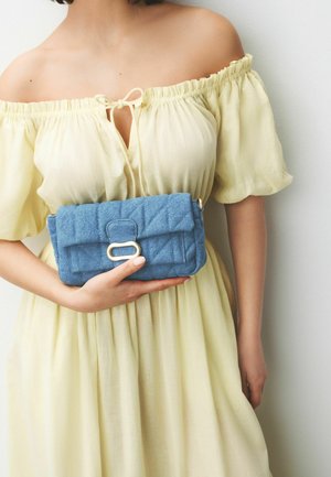 Nő halvány sárga váll nélküli ruhában, aki egy kis, steppelt kék táskát tart aranyszínű csattal.