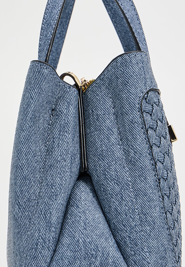 Sac en denim bleu clair, arbore une texture tissée unique sur un côté, une finition lisse, et une fermeture éclair dorée en haut.