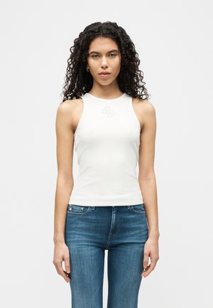 Giovane donna con capelli lunghi e ricci, indossa una canotta bianca senza maniche e jeans blu, in piedi contro uno sfondo chiaro e uniforme.