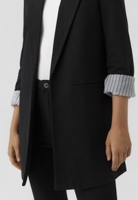 Mustan pitkä bleiseri, jossa on muotoiltu leikkaus, yksi rintatasku ja ylöskäännetyt hihat harmaavalkoisella raidallisella kuviolla.