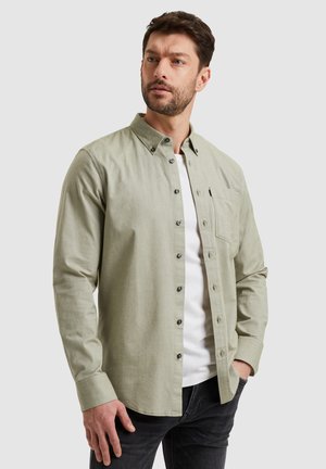 Camicia button-up in tessuto verde chiaro con maniche lunghe, colletto, taschino sul petto e bottoni scuri. Indossata su una maglietta bianca.