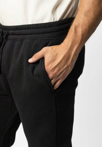 Zwarte sweatpants met een elastische tailleband en zijzakken. Het materiaal lijkt zacht en gestructureerd. Hand rust in de rechterzak.