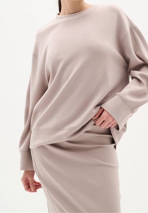 Kobieta nosząca luźną beżową bluzę i dopasowaną spódnicę, z jedną ręką delikatnie wsuniętą w pas spódnicy.