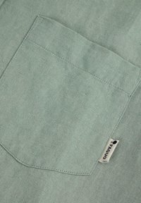 Texturiertes mintgrünes Material mit einer genähten Tasche und einem kleinen Etikett mit der Aufschrift "FAGUO." Der Stoff hat eine weiche, matte Oberfläche.