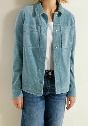 Kvinde iført en teal kordfløjlsjakke med knapper over en hvid skjorte og blå jeans, stående foran en neutral baggrund.