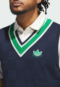 Mørkeblå strikket golfvest med v-hals, grønne og hvide striber i kanten og et emblem med et bladlogo og teksten "GOLF KLUB".
