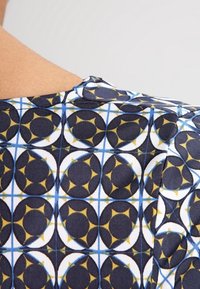 Tričko s krátkým rukávem s geometrickým vzorem obsahujícím kulaté tvary v modré, černé a žluté barvě na bílém pozadí. Měkká, hladká textura.