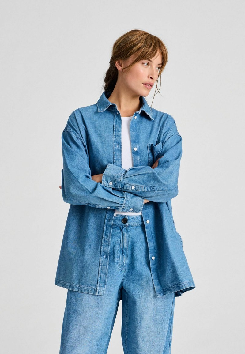Lyseblå denimskjorte med knaplukning, lange ærmer, brystlomme og afslappet pasform, kombineret med matchende denimbukser. Glat tekstur.