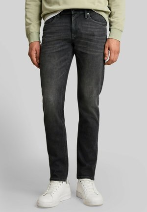 Mann trägt ausgewaschene schwarze Jeans, weiße Sneakers und ein hellgrünes Sweatshirt, steht vor einem schlichten grauen Hintergrund.