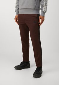 Barna sportnadrág fekete oldalsó csíkokkal, bal combján Adidas logóval, puha anyagból készült, fekete cipőkkel párosítva.