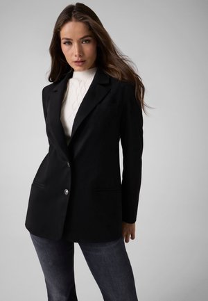 Mujer con cabello largo y castaño que lleva una blazer negra sobre un suéter de cuello alto blanco texturizado y jeans oscuros, de pie frente a un fondo gris liso.