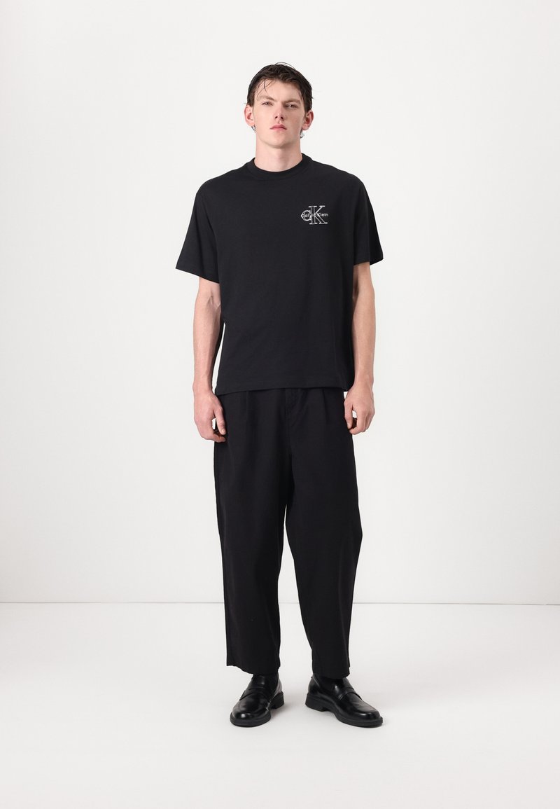 Black cotton t-shirt with a printed logo on the chest, paired with loose-fitting black trousers and black leather shoes.