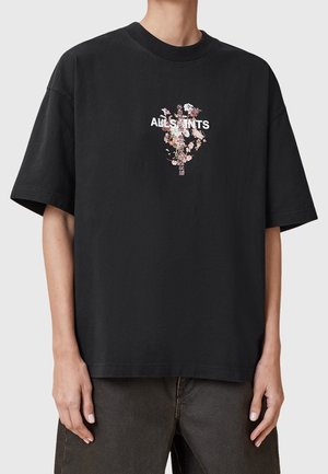 Osoba nosząca czarny, oversize'owy t-shirt z kwiatową grafiką i napisem "ALLSAINTS" na piersi, w połączeniu z ciemnymi spodniami.