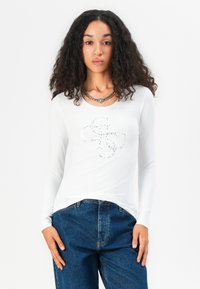 Langärmliges weißes T-Shirt mit einem dekorativen silbernen Strassmuster auf der Vorderseite, kombiniert mit hoch taillierten blauen Jeans.