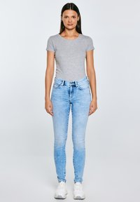 Naine seisab näoga ettepoole, seljas halli lõikega t-särk, helesinised kitsad teksad ja valged tossud, ühtlase taustaga.