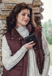 Schwarze Lederbrieftasche mit Klappenverschluss und eingeprägtem Logo, gehalten von einer Person, die einen grob gestrickten hellbeigen Pullover und eine burgunderfarbene Steppweste trägt.