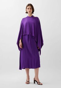 Violetti mekko leveillä hihoilla, virtaavalla kankaalla, polvipituisella suunnittelulla ja korkealla kaula-aukolla. Yhdistetty mustiin remmisandaaleihin.