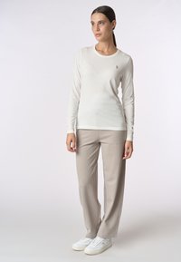 Camicia a maniche lunghe color crema in tessuto leggero, con scollatura rotonda e un piccolo logo. Abbinata a pantaloni beige dalla vestibilità ampia e sneakers bianche.