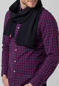 Bufanda de punto azul marina colgada alrededor del cuello de una persona, combinada con una camisa a cuadros roja y azul. Textura suave, diseño simple y etiqueta visible.