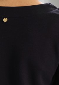 Top de punto negro con escote redondo y un detalle metálico dorado en la parte posterior. Textura suave y diseño ajustado.