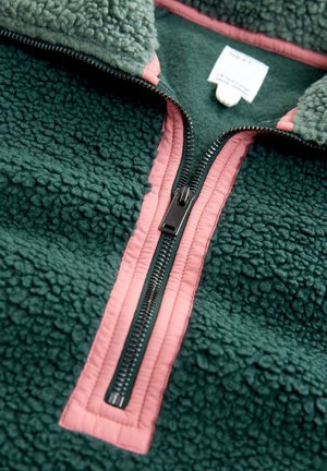 Žalias vilnonis džemperis su aukštu apykaklės kraštu, kontrastingo rožinio užtrauktuko ir briaunos detalėmis, tekstūruotu paviršiumi ir etiketė matoma netoli apykaklės.