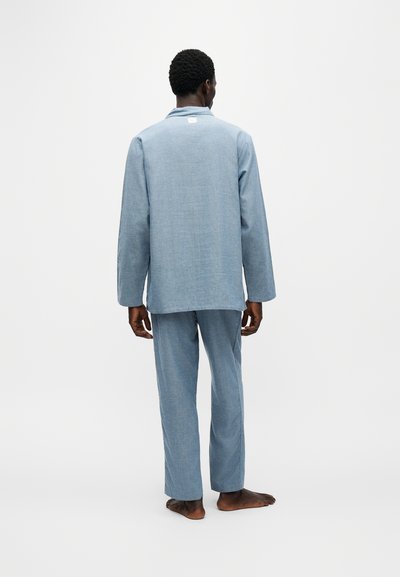 Homme debout pieds nus, dos tourné, portant un haut ample bleu clair à manches longues et un pantalon assorti sur un fond blanc uni.
