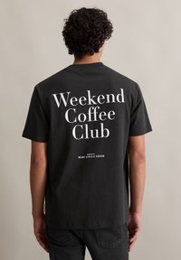 Zwarte katoenen t-shirt met korte mouwen, voorzien van grote witte tekst op de achterkant: "Weekend Coffee Club" met "georganiseerd door Marc O'Polo Denim."