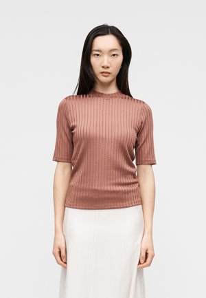 Vrouw met stijl zwart haar draagt een geribd, kort mouw, roestkleurig topje en een witte rok, staande tegen een effen lichtgrijze achtergrond.