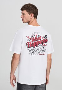 Biały bawełniany t-shirt z graficznym nadrukiem kart do gry i kości na plecach. Napis brzmi "Co się dzieje w VEGAS." Krótkie rękawy.