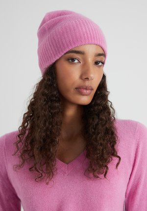 Donna con capelli ricci e scuri che indossa un berretto di lana rosa e un maglione rosa a scollo a V, su uno sfondo grigio uniforme.