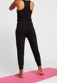 Débardeur noir de sport et pantalon en tissu doux et extensible. Le pantalon présente une taille élastique et des chevilles côtelées, placé sur un tapis rose.