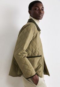 Olive green quilted jacket with a zip front, ribbed collar, and side pockets. Features decorative stitching and a relaxed fit.