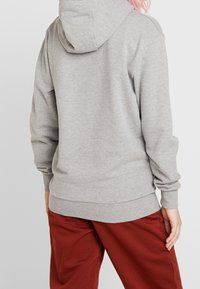 Felpa grigia con cappuccio, dal tessuto morbido, con polsini e orlo a coste. Presenta una tasca a marsupio e dettagli di cucitura, indossata con pantaloni bordeaux.