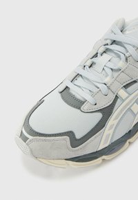 Sneakers azul claro e cinza com materiais de malha e camurça, camadas intrincadas, ponta arredondada e uma sola acolchoada e texturizada.