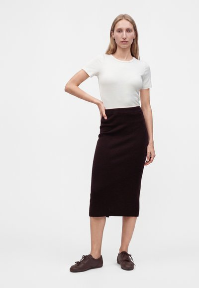 Femme aux longs cheveux blonds portant une chemise blanche à manches courtes, une jupe midi noire et des chaussures marron, debout avec une main sur la hanche.