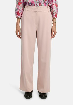 Person iført løstsiddende blege lyserøde bukser, sorte sko og en blomsterlangærmet skjorte, stående med armene afslappet ned langs siderne.