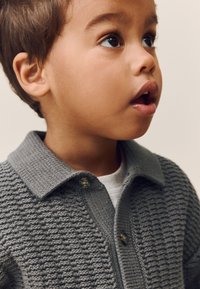Gray knitted polo shirt with a textured pattern, two buttons at the collar, and a white undershirt visible beneath.
