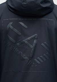 Μαύρο μπουφάν με κουκούλα με ανάγλυφο λογότυπο Giorgio Armani και στυλιζαρισμένα γράμματα στην πλάτη, μέσα σε τετράγωνο πλαίσιο με ραφή.