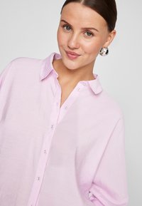 Lys rosa skjorte med knappestolpe, myk tekstur, krage og løs passform. Har knapper foran og lett oppbrettede ermer.