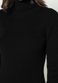 Pull noir à col roulé côtelé en tissu doux. Présente une coupe ajustée avec un logo subtil sur le col.