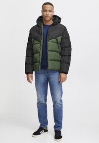 Pufjakke med aftagelig hætte, sort overdel og grøn krop, lynlås lukning, kviltet tekstur, parret med blå jeans og sorte sneakers.