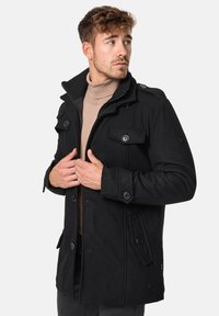 Abrigo negro de mezcla de lana con diseño de doble botonadura, grandes botones y bolsillos frontales. Cuello alto y hombros estructurados.